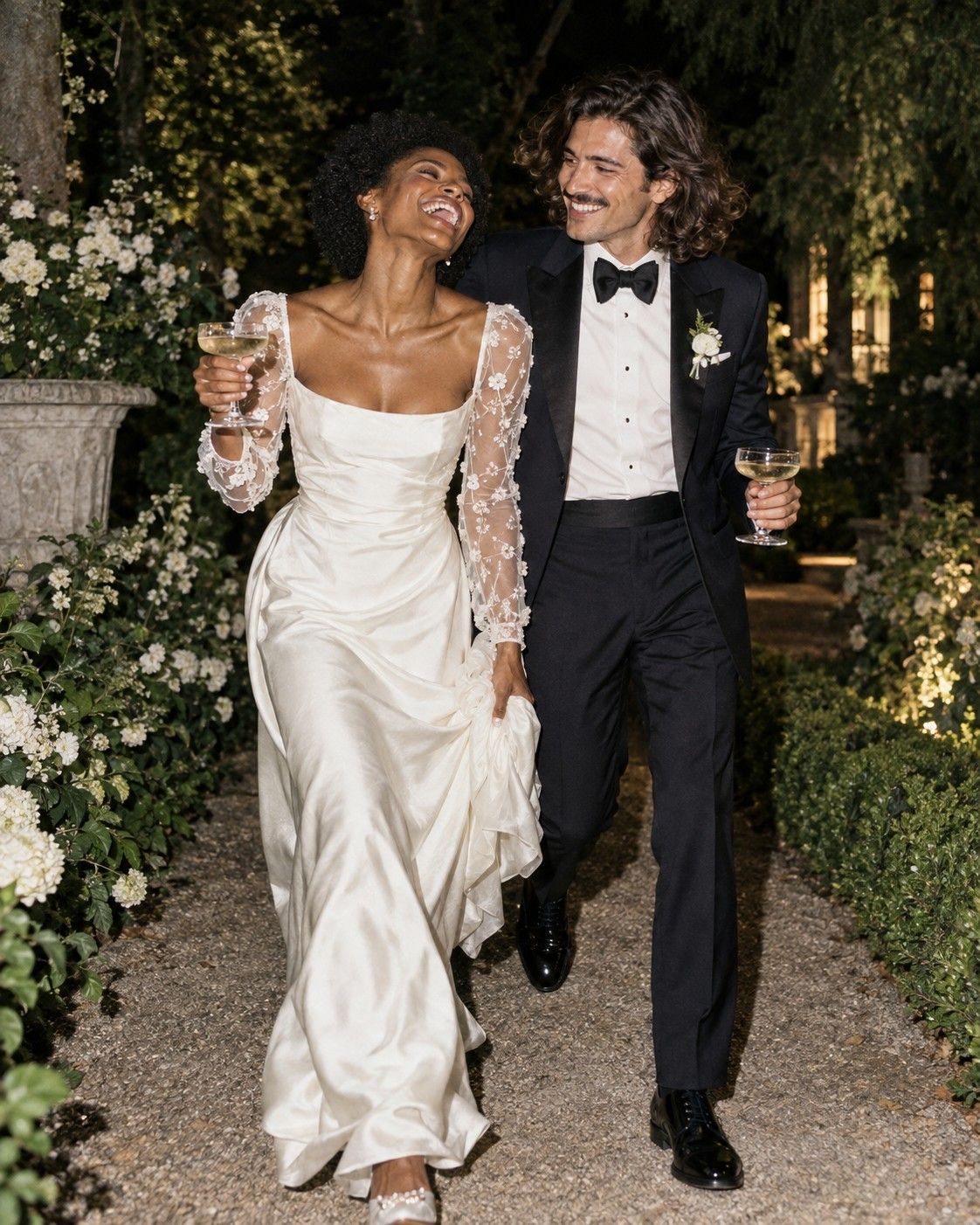 A bride laughing and a groom smiling at her as they walk along a garden path at night, holding champagne glasses.