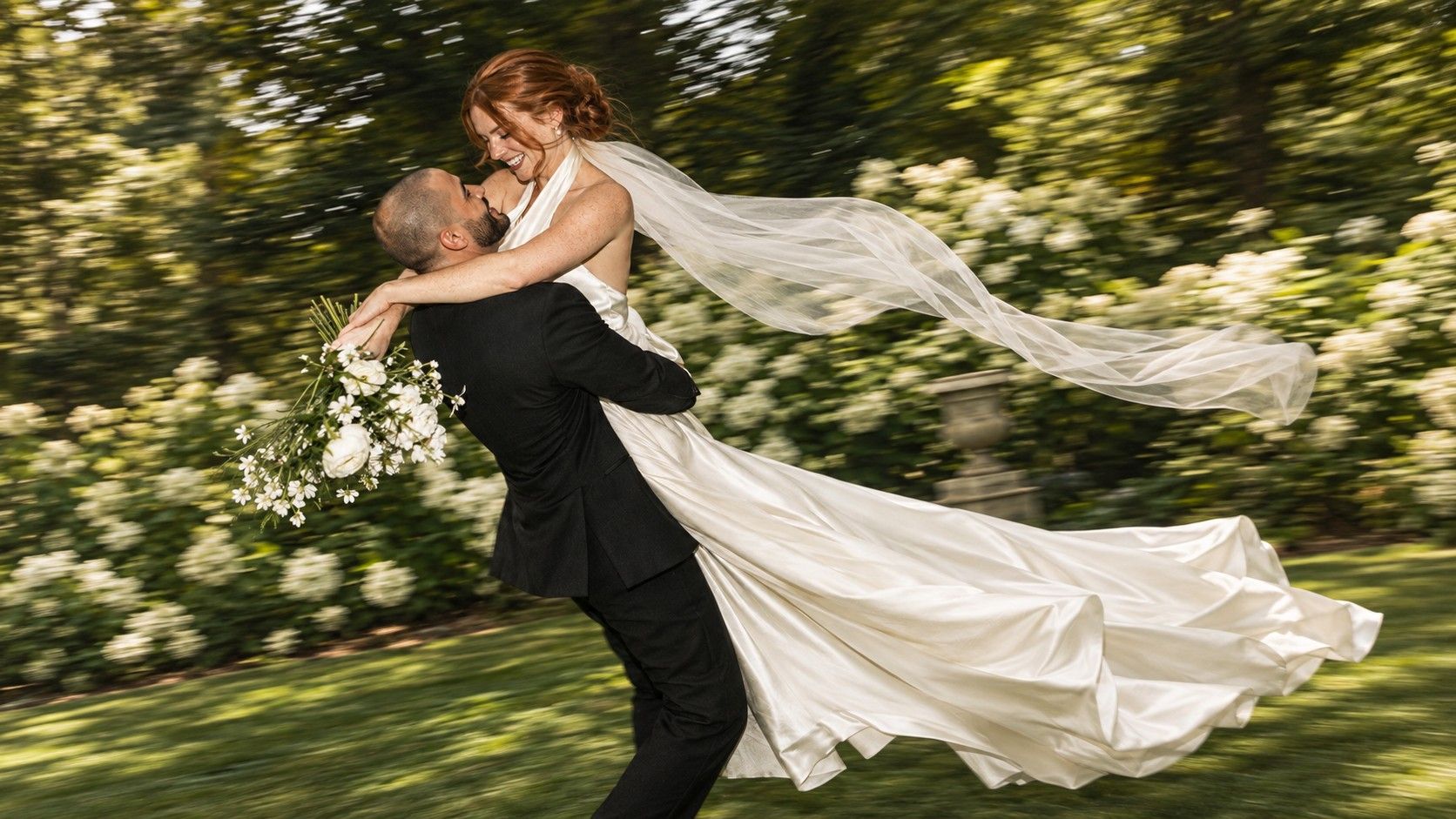 A groom lifting and spinning a laughing bride in a garden, her veil flying.